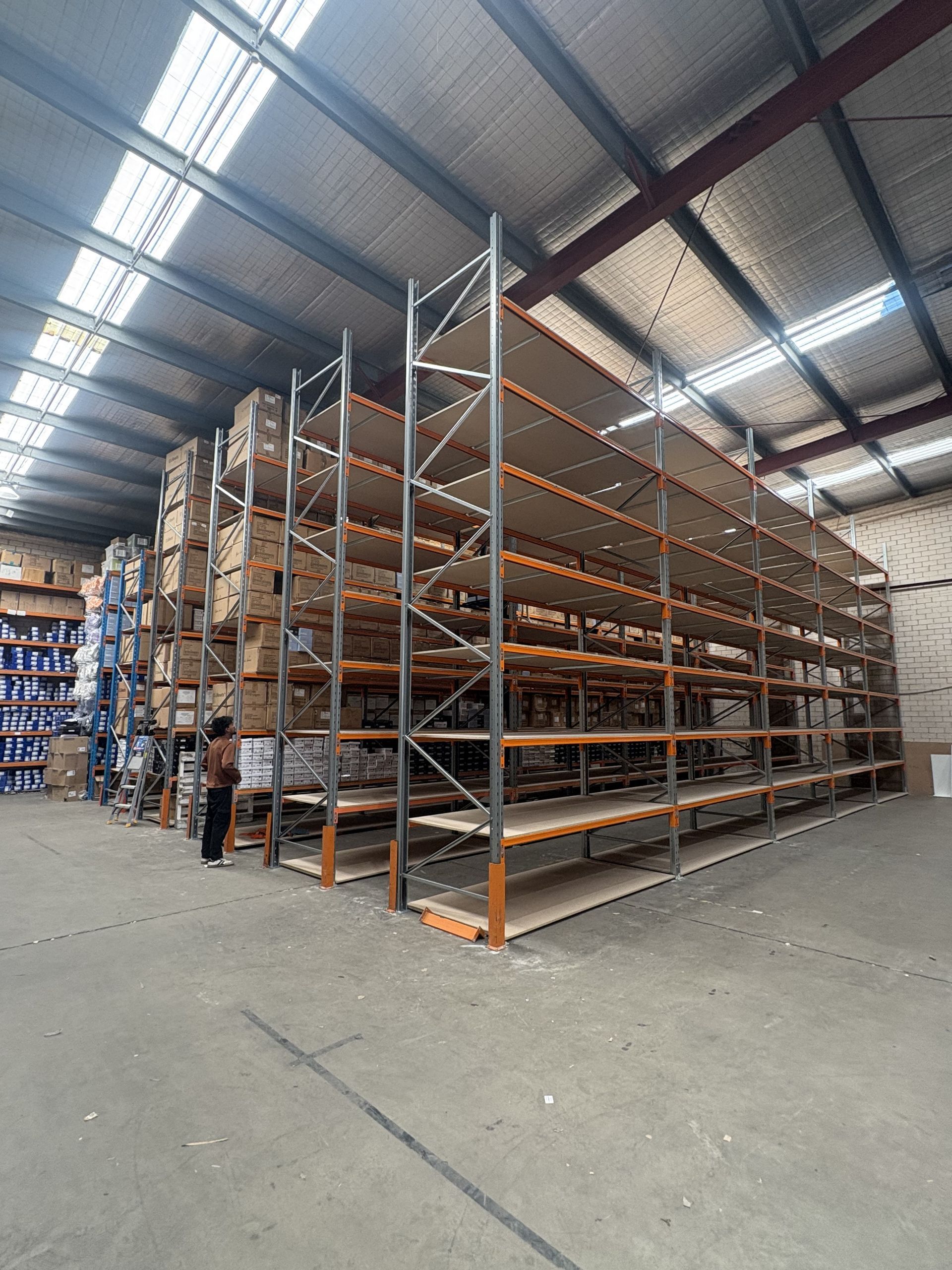 Warehouse aisle with tall metal shelving stacked with boxes and pallets under bright skylights — Shop Fittings Office Furniture Materials Handling In Gosford, NSW