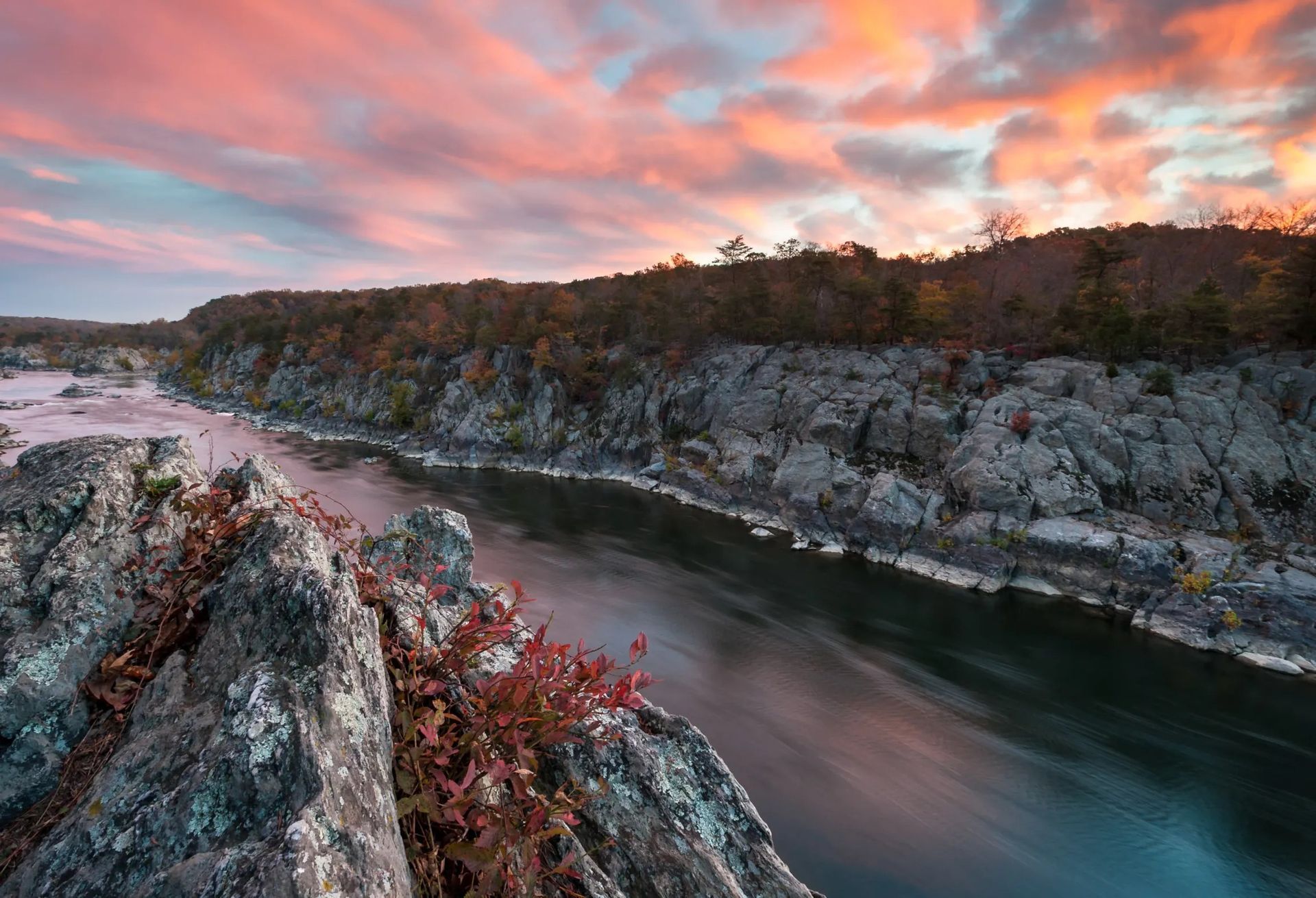 A rocky river gorge at dawn or dusk, with reddish autumn foliage clinging to stone in the foreground and a wide sky streaked with pink and orange clouds above tree-lined cliffs. The scene conveys quiet vastness and the turning of seasons, a place of natural stillness rather than any built setting.