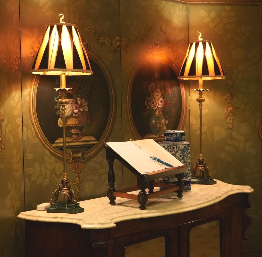 A similar antique console setup features an open guestbook and pen flanked by two brass lamps with striped shades, set before oval mirrors and painted floral panels on a gold wallpapered screen. The soft golden lighting and inviting blank book suggest an intimate space meant for quiet reflection or leaving written words.
