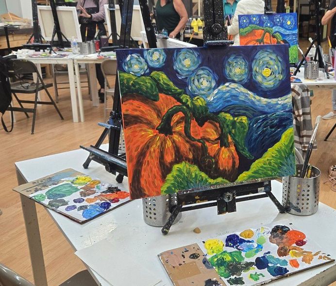 Paintings on easels at a craft fair, including a colorful pumpkin scene and paint palettes on tables