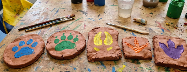 Painted clay paw-print and cat-footprint tiles drying on a worktable with brushes and paint jars nearby