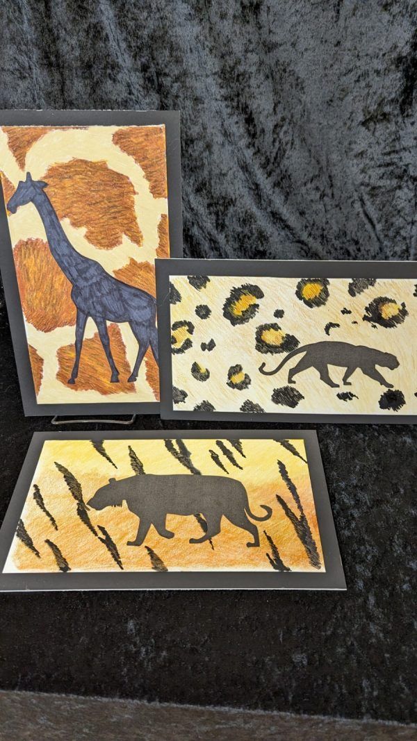 Three framed animal prints leaning on a dark fabric surface, with giraffe, leopard, and elephant designs.