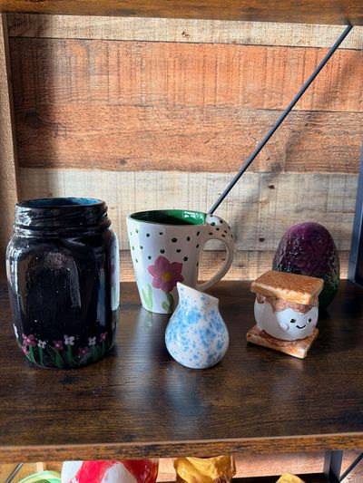 Assorted handmade pottery pieces and a black jar on a wooden shelf against a rustic wall.