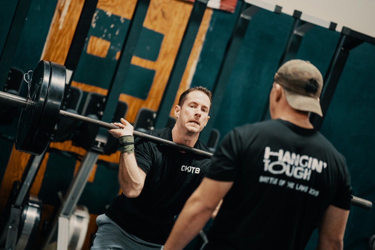 Man lifting barbell in gym, aided by a spotter wearing a
