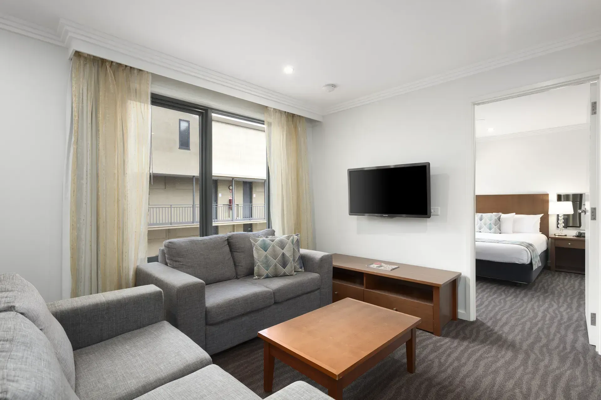 Modern living room with gray sofas, wooden coffee table, wall-mounted TV, and open bedroom doorway