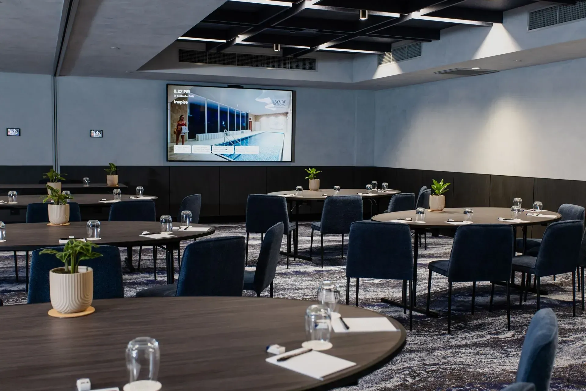 Empty conference room with round tables, dark chairs, and a wall-mounted presentation screen.