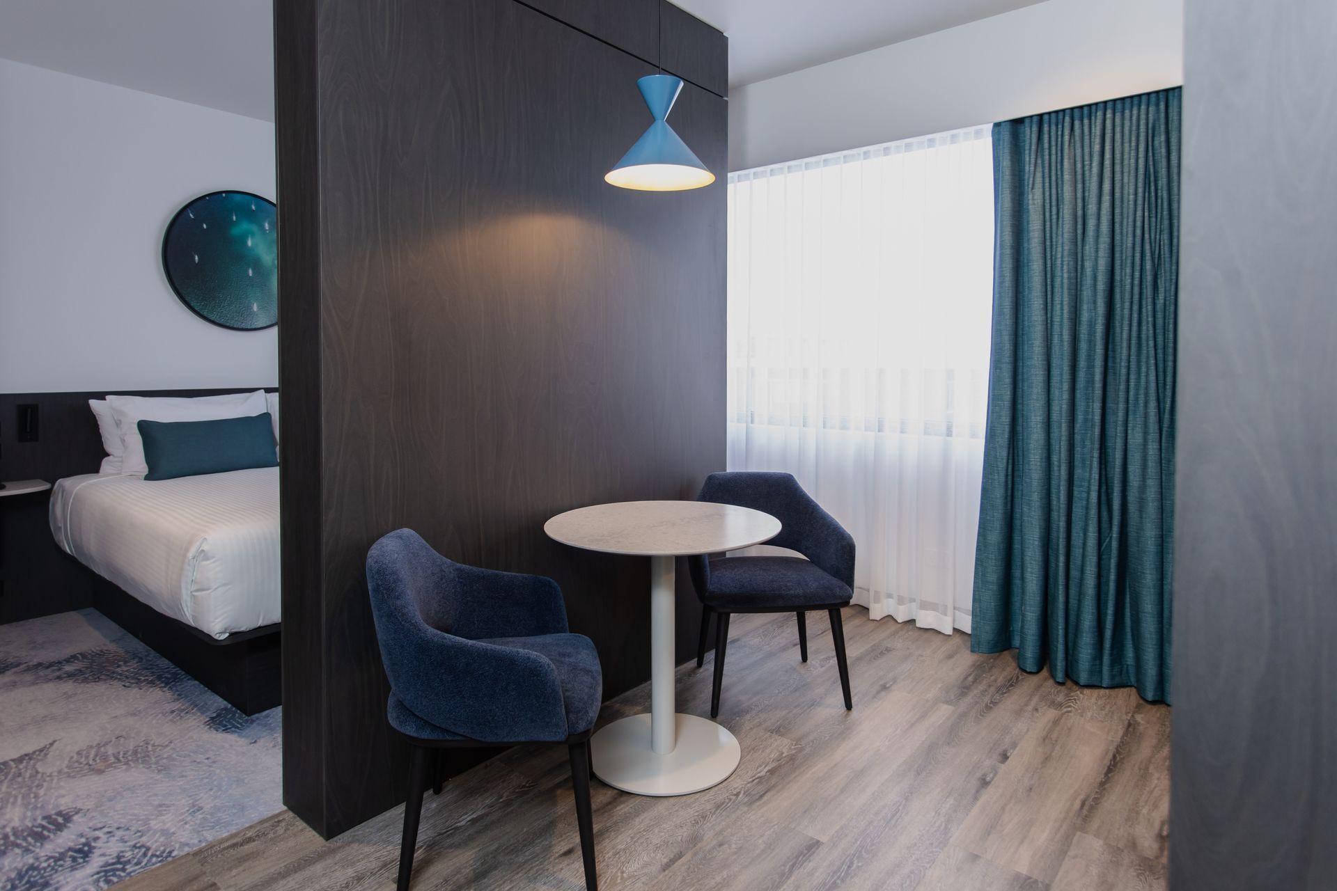 Modern hotel room with blue chairs, round table, bed, and sheer curtains by a window