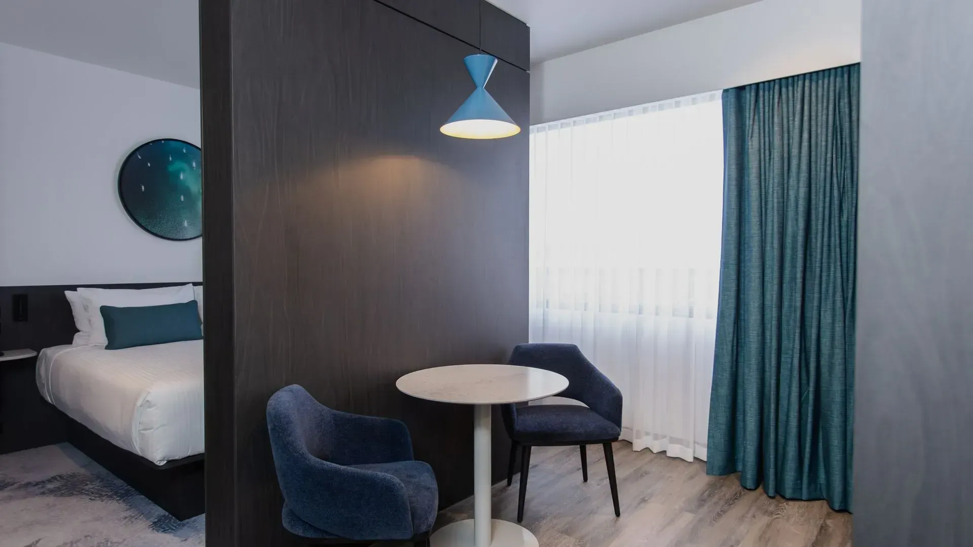 Modern hotel room with blue chair, small round table, bed, and large window with curtains