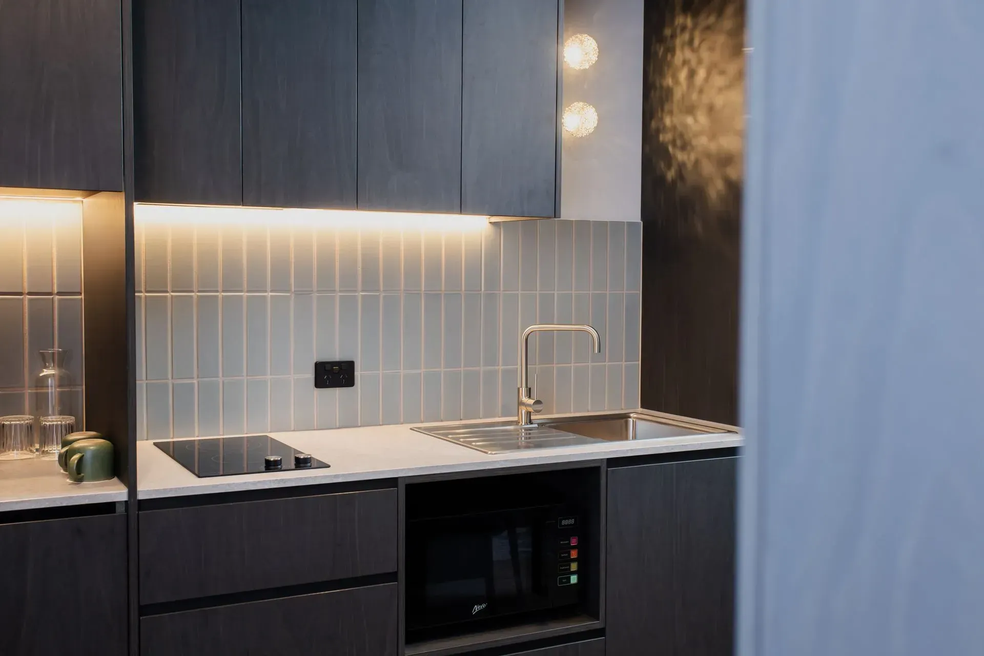 Modern kitchen with dark cabinets, white tile backsplash, sink, and built-in oven under under-cabinet lighting