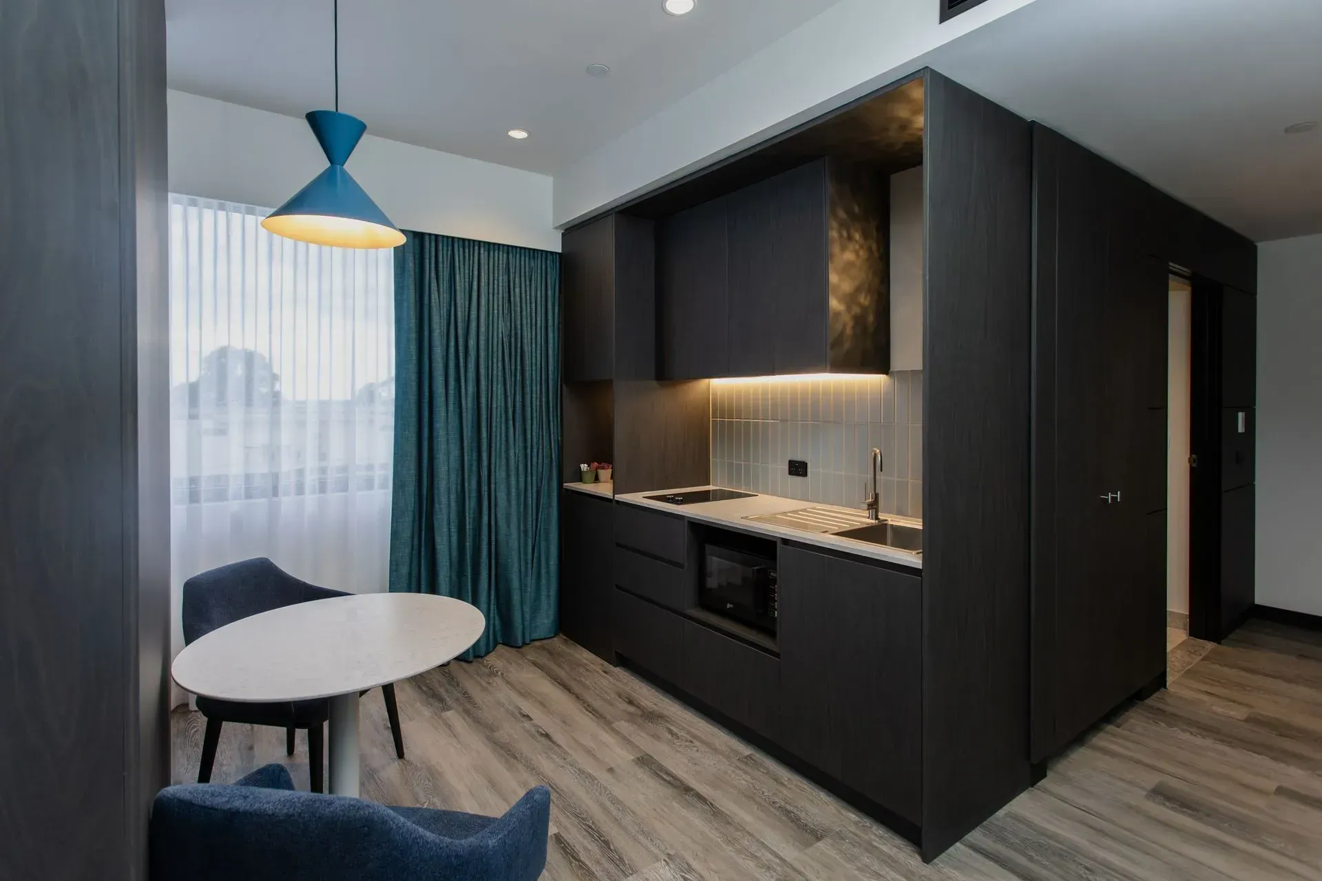 Modern studio apartment kitchen nook with dark cabinets, round table, blue curtains, and wood flooring