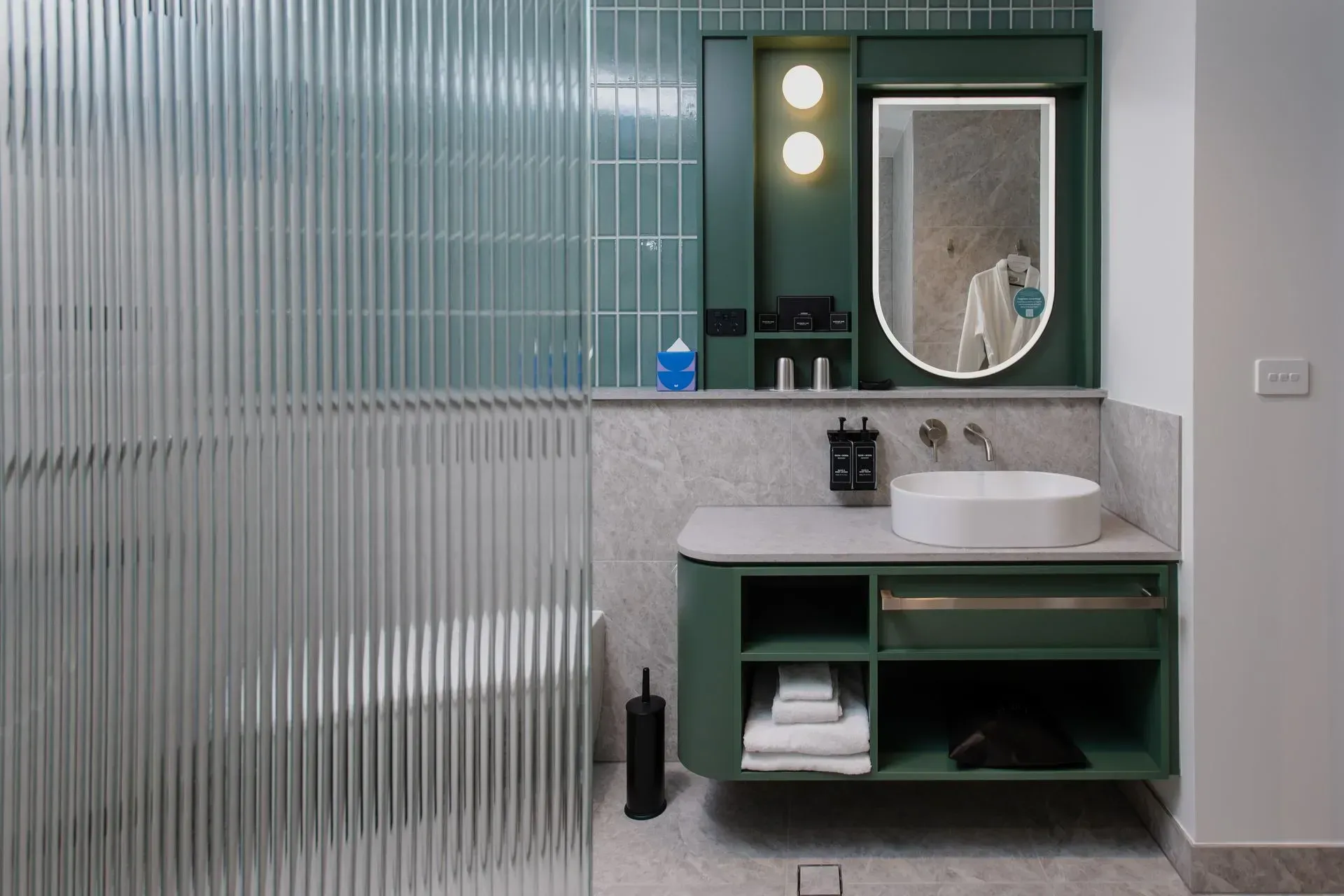 Modern bathroom vanity with green cabinets, round mirror, white sink, and frosted glass shower wall