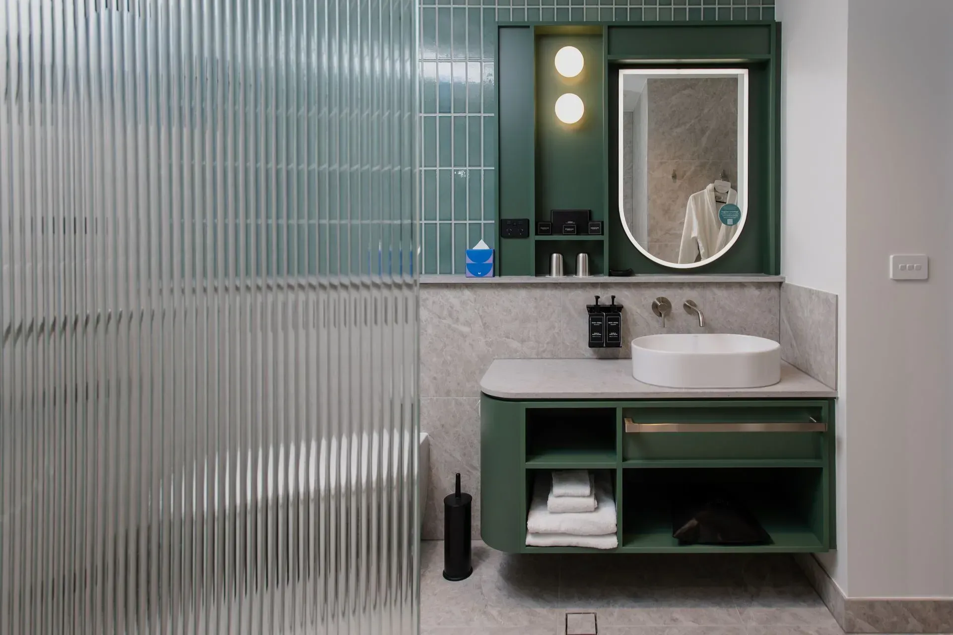 Modern bathroom vanity with green cabinets, round mirror, sink, and frosted glass shower partition