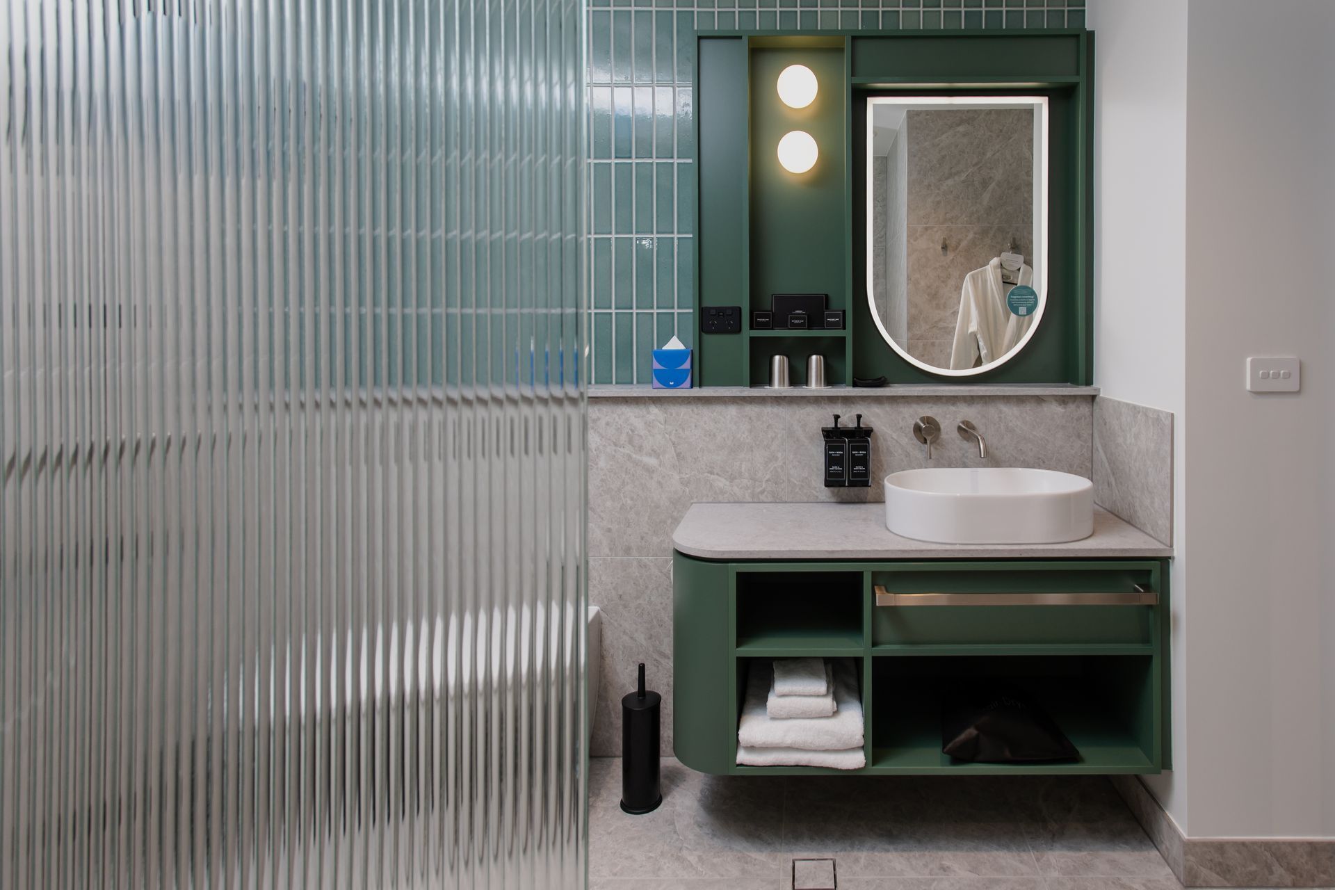 Green vanity with round mirror and white sink in a tiled bathroom
