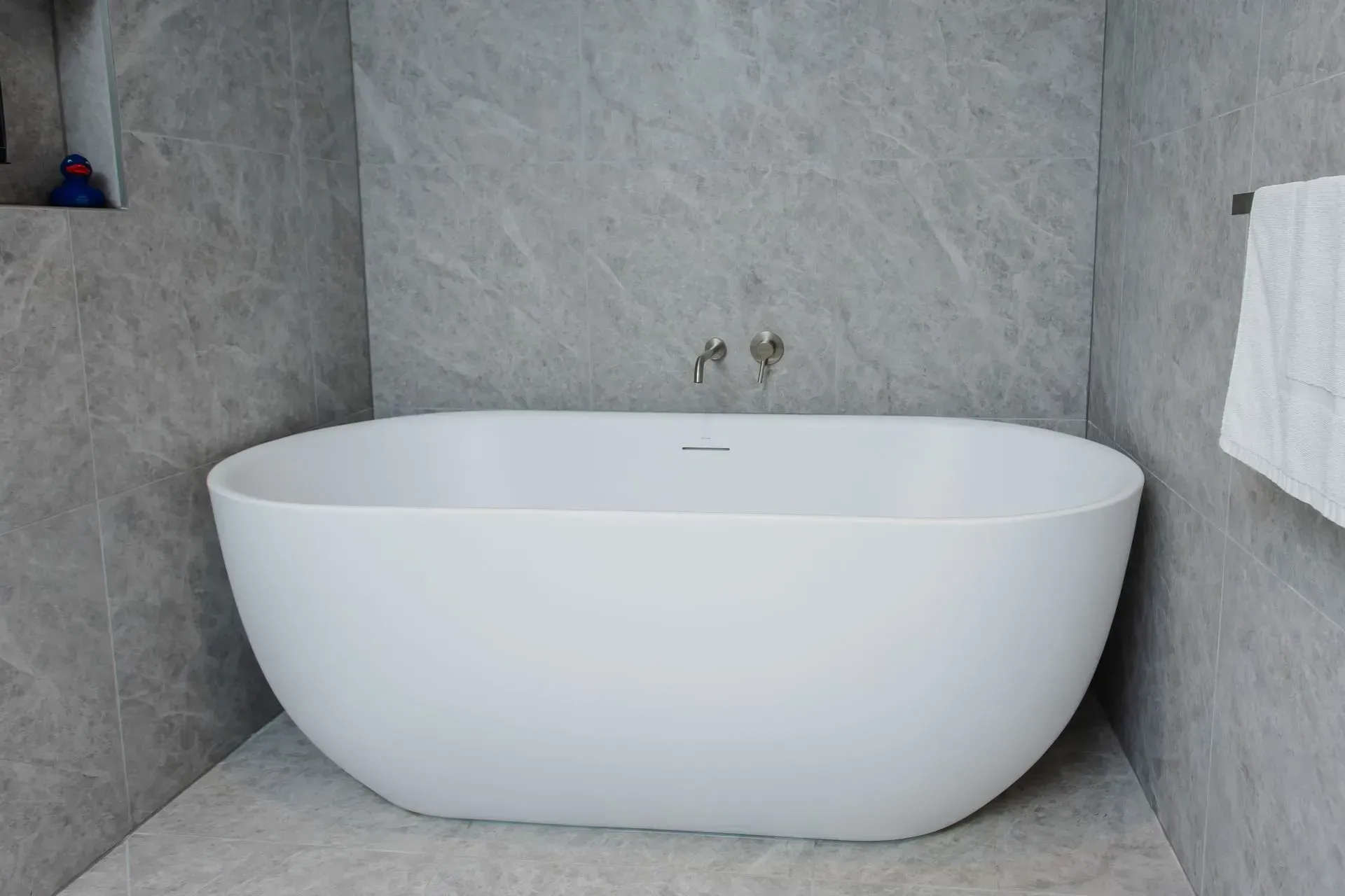 Modern white freestanding bathtub in a gray tiled bathroom alcove