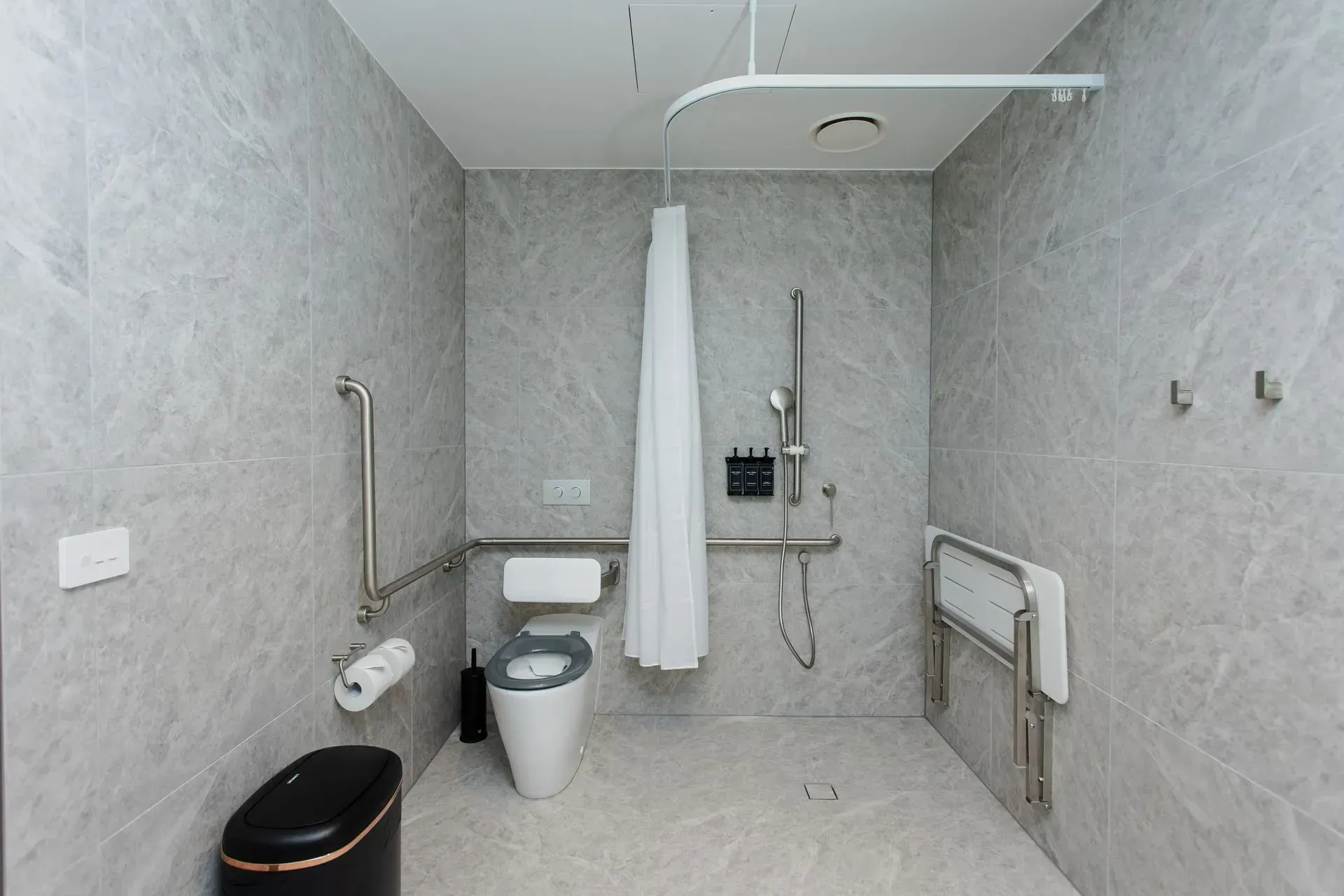 Accessible bathroom with toilet, shower, grab bars, and fold-down seat on white tiled floor.