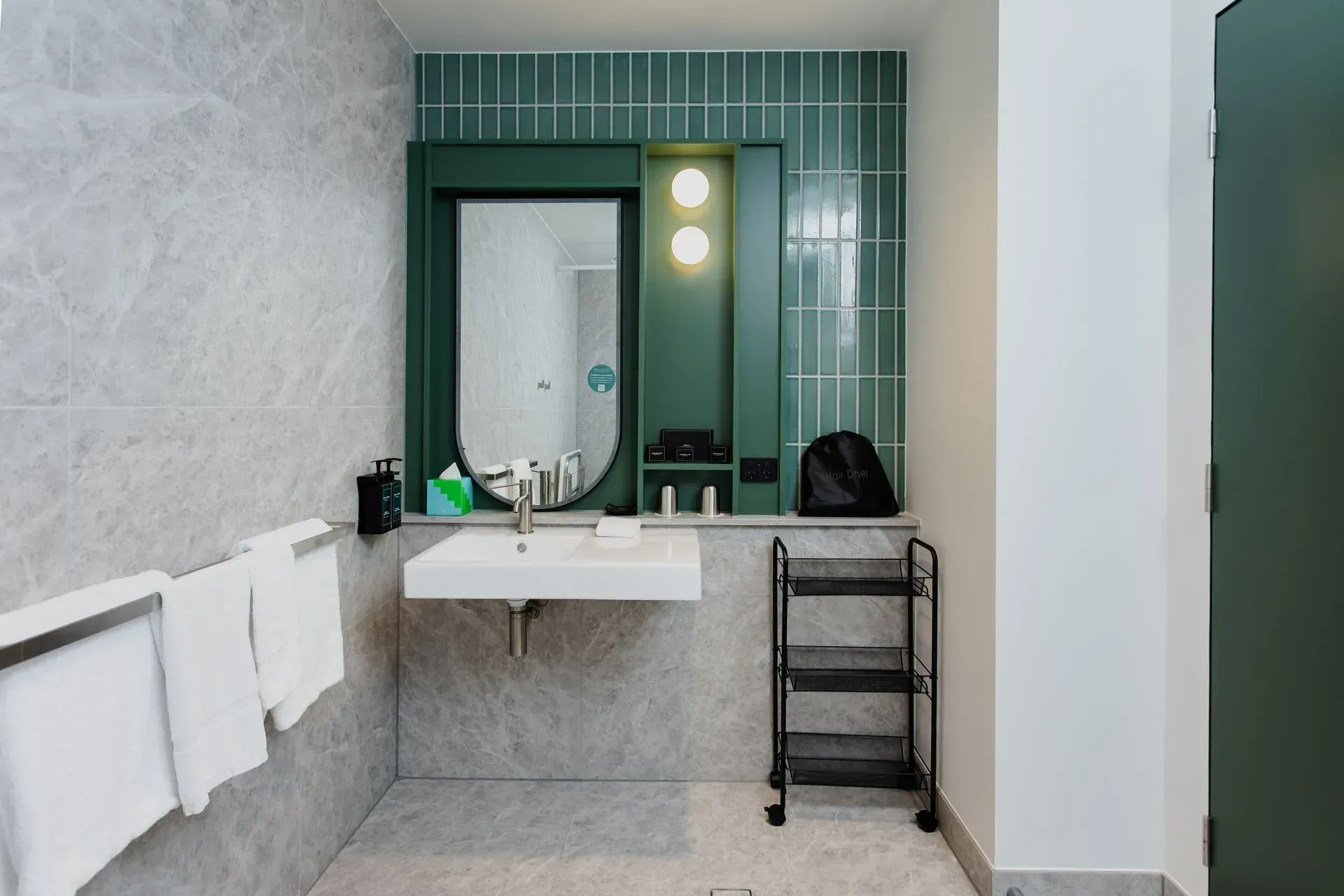 Modern bathroom with green-tiled vanity wall, double sink, mirror lights, towels, and a black cart