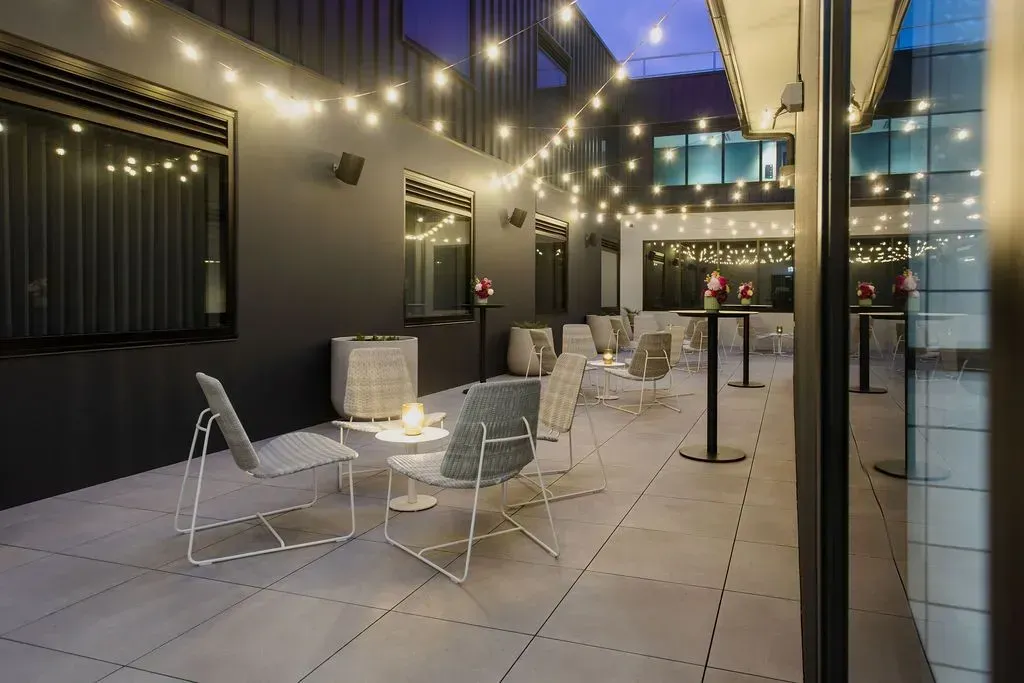 Outdoor patio with string lights, lounge chairs, and tables beside modern buildings at dusk