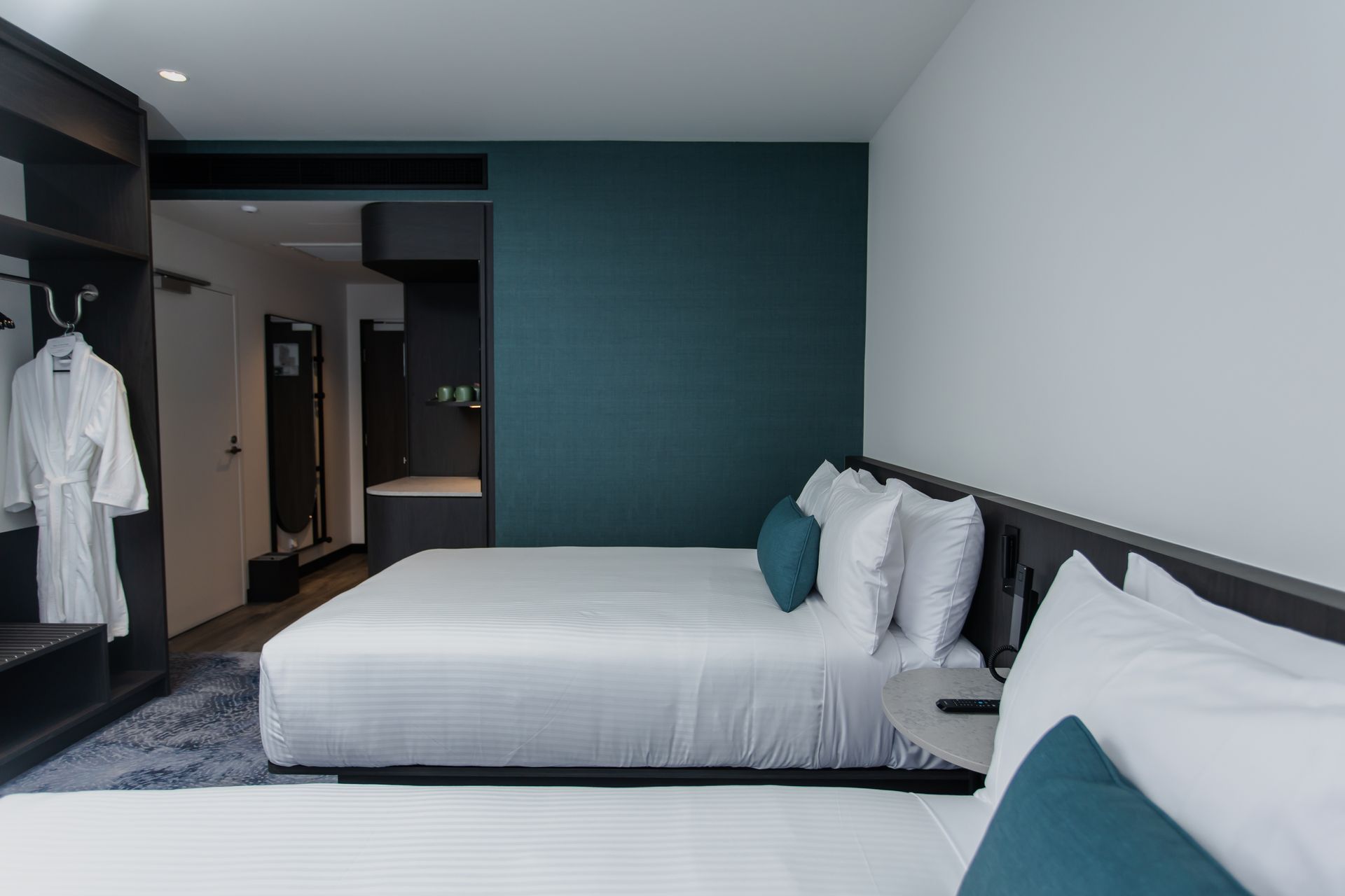 Modern hotel room with two beds, teal accent wall, open closet, and hallway view