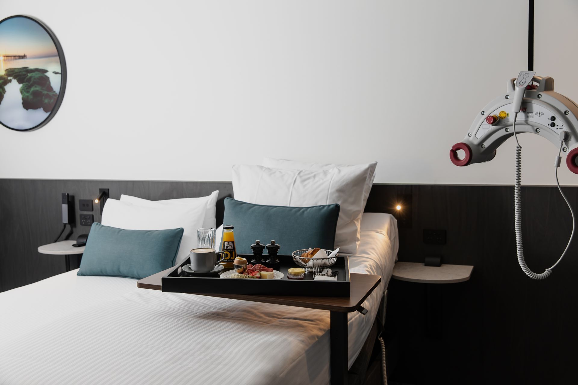 Modern hotel room with neatly made bed, breakfast tray, and hanging assist lift equipment beside the bed