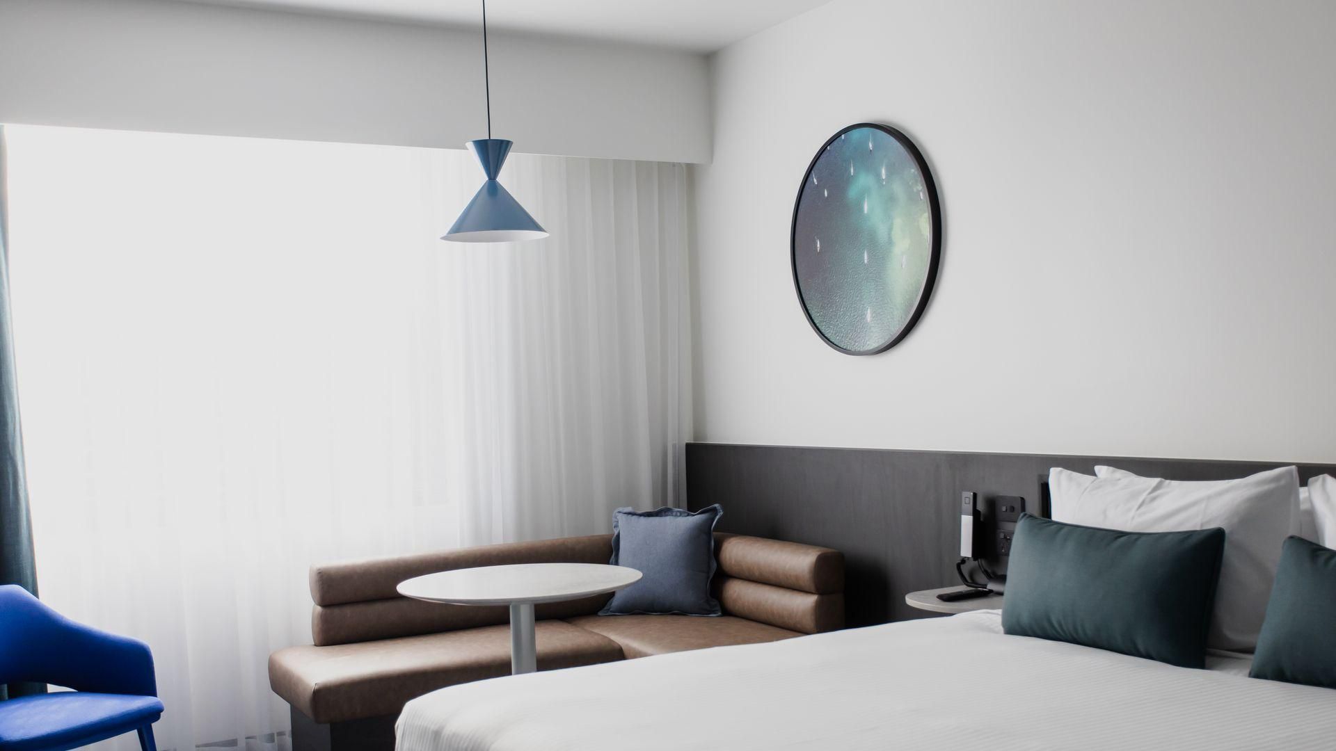 Modern hotel room with bed, blue chair, round table, sofa, and large window with sheer curtains.