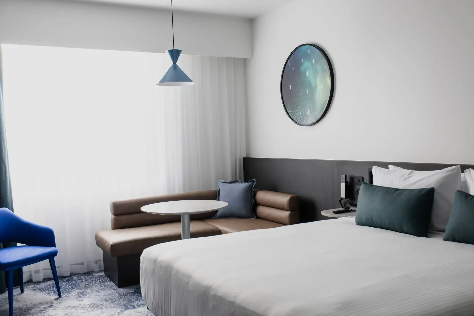 Modern hotel room with a bed, sofa, blue chair, small table, and large window with white curtains.