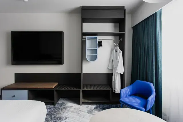 Modern hotel room with wall-mounted TV, desk, wardrobe, blue chair, and teal curtains