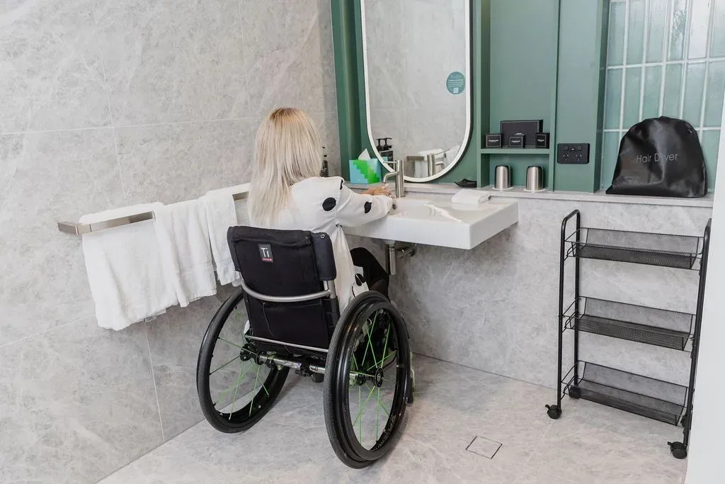 Person in wheelchair at bathroom sink, with towel and mirror in a bright accessible bathroom.