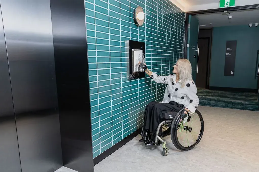 Woman in wheelchair using a wall-mounted drink station in a hallway with teal tiled wall