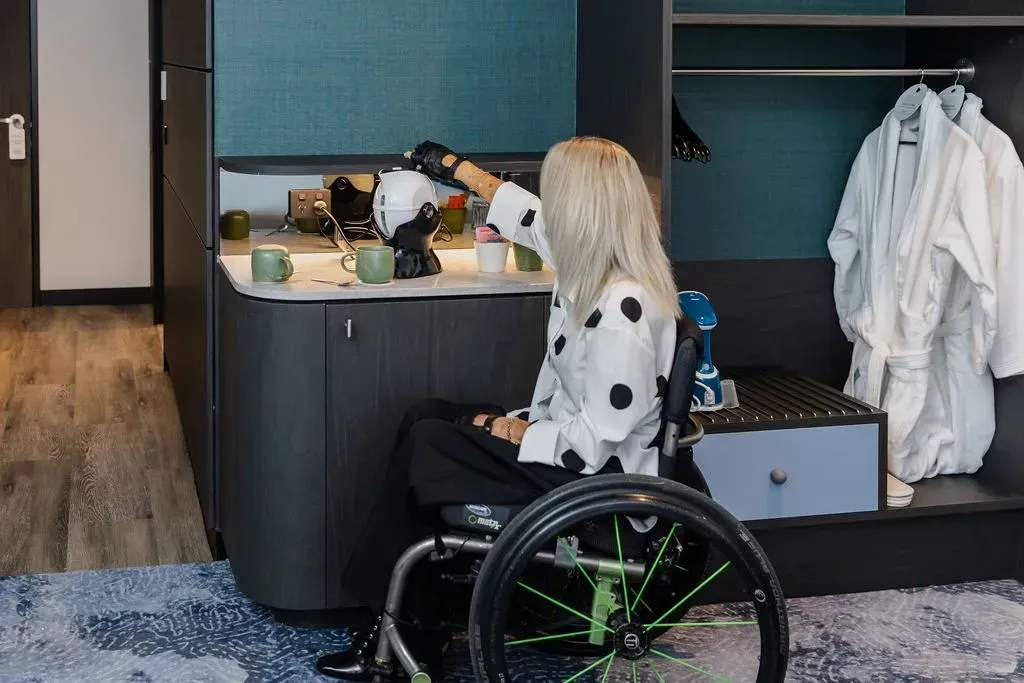 Person in a wheelchair at a vanity in a modern room, with hanging white garments nearby.