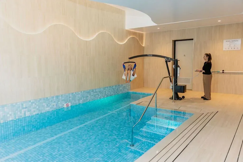 Indoor pool with blue water, tiled deck, and a pool lift beside a woman standing at the edge