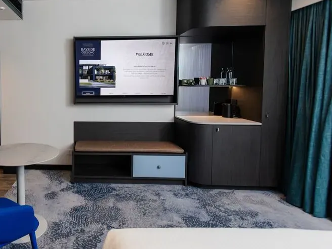 Hotel room seating area with wall-mounted TV, bench, small table, and built-in shelf