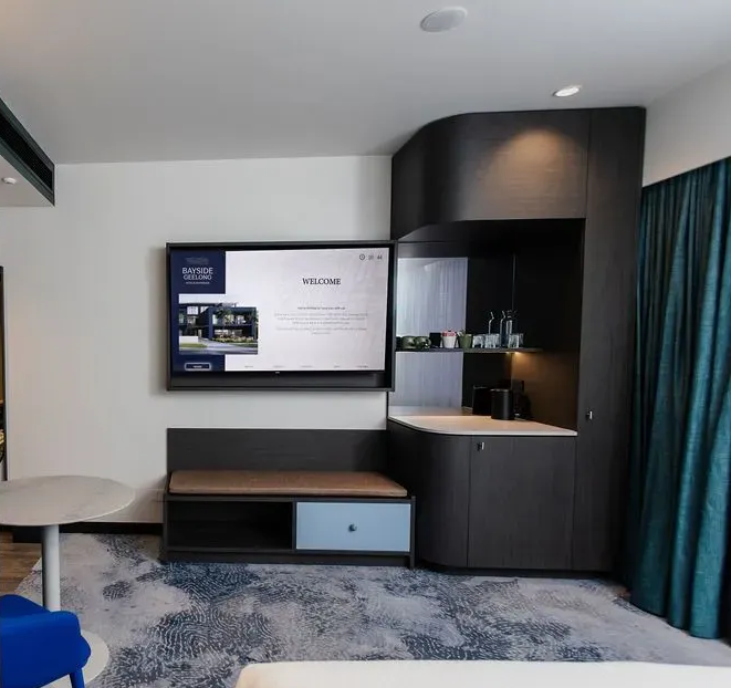 Modern hotel room with wall-mounted TV, built-in desk, small table, and teal curtains