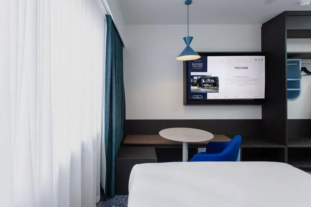 Modern hotel room with a bed, desk, blue chair, wall-mounted TV, and sheer curtains beside a window