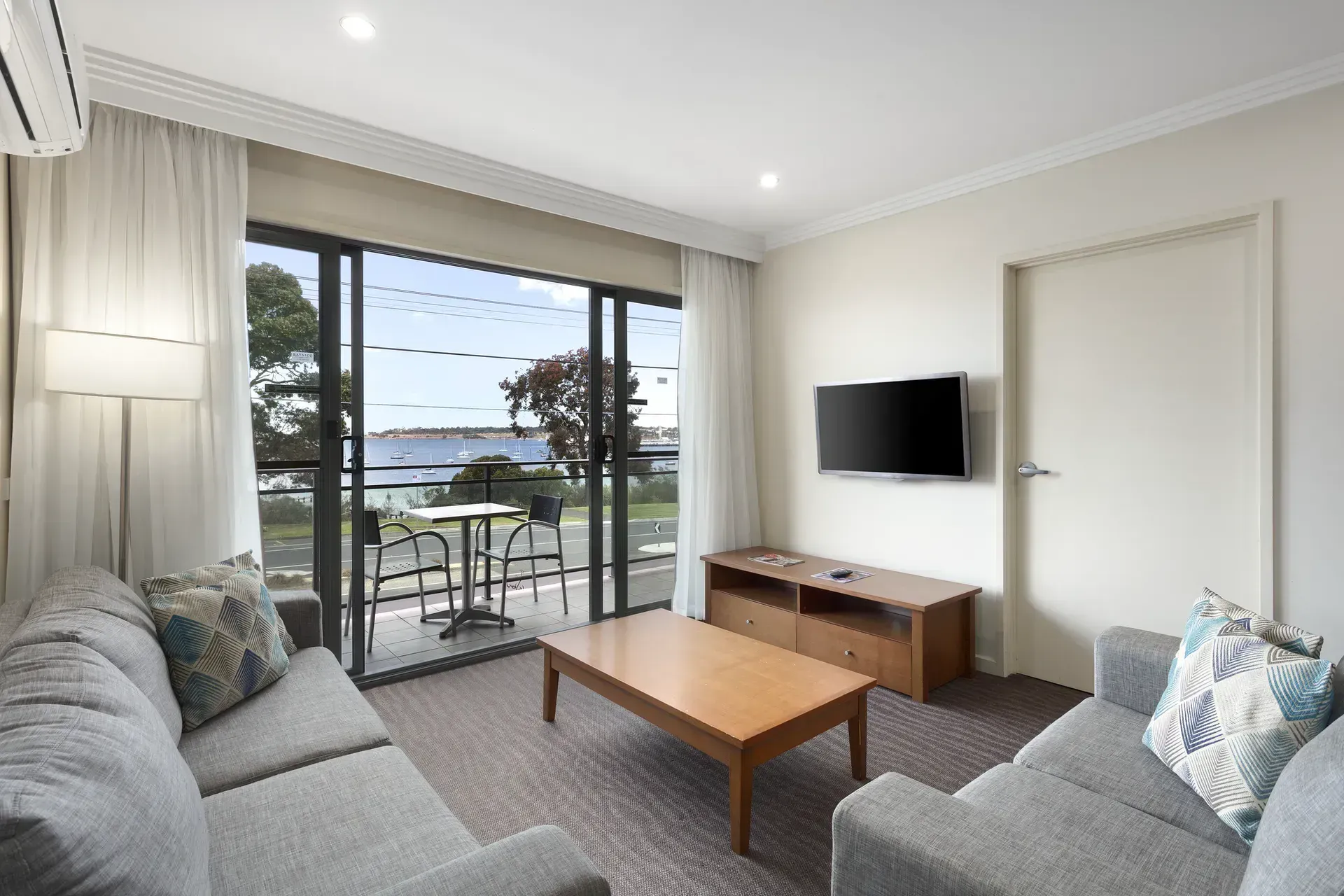 Bright living room with gray sofas, wall-mounted TV, and balcony doors overlooking the water