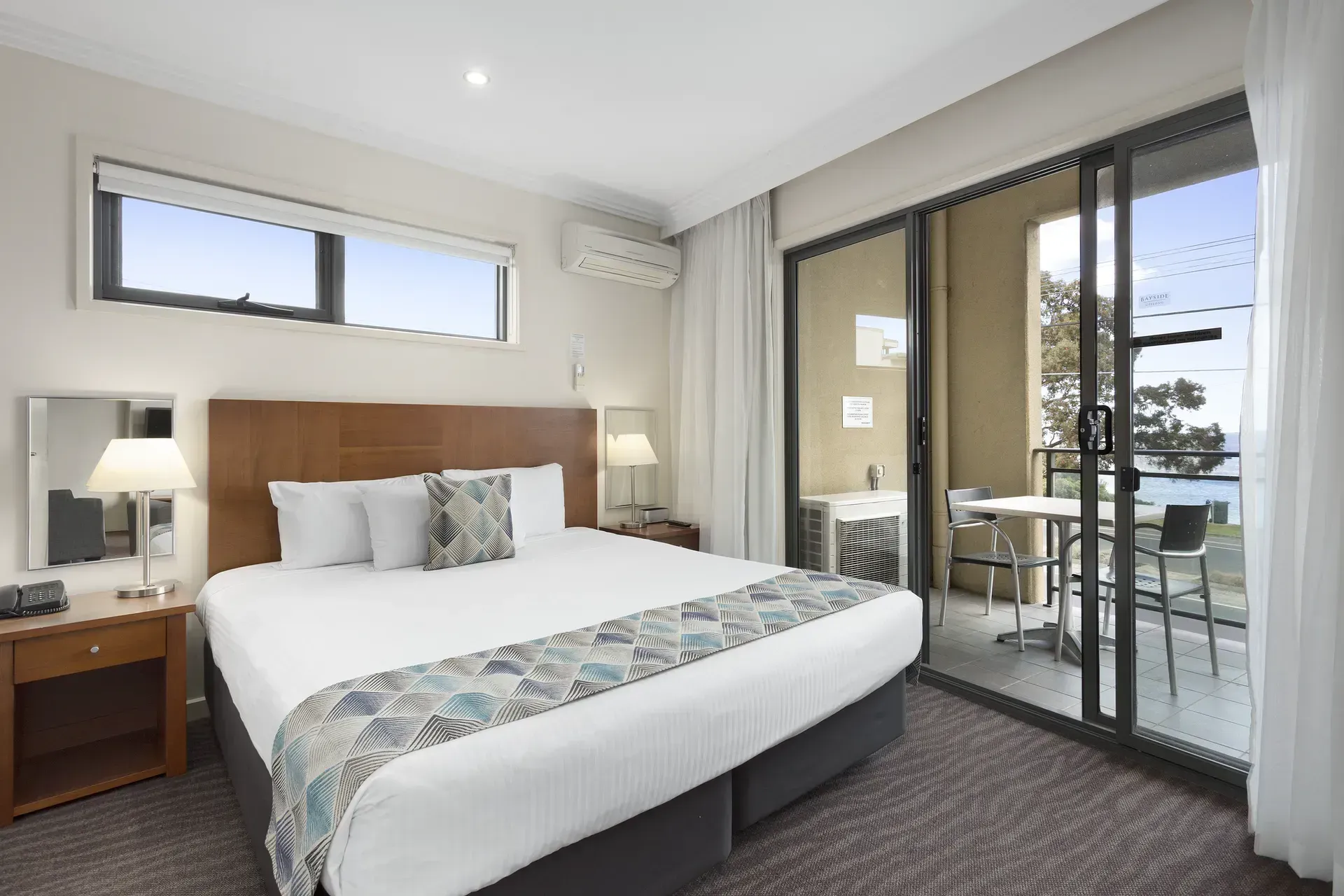Modern hotel bedroom with king bed, nightstands, and sliding glass doors to a balcony overlooking water