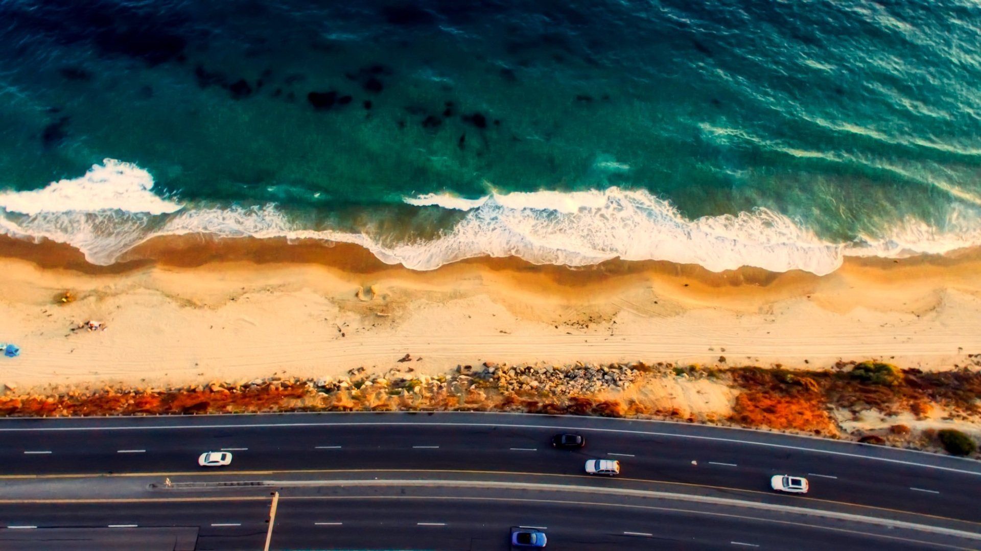 Aerial view of turquoise waves washing onto a sandy beach beside a coastal road with cars.