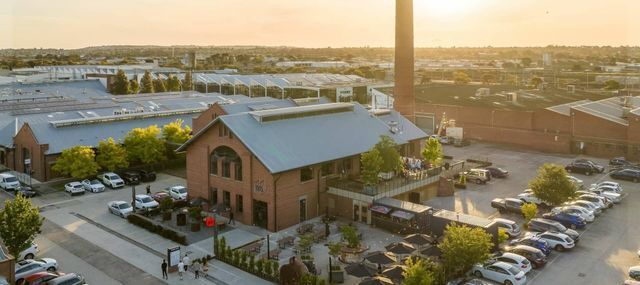 Aerial view of a brick building beside water at sunset, with parked cars and a tall chimney nearby