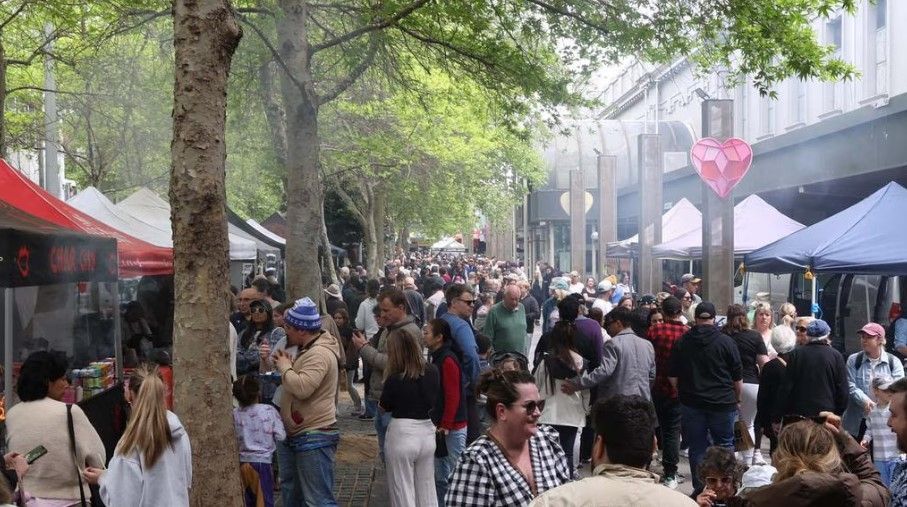 Crowded outdoor market or festival walkway with tents, trees, and people browsing in daylight