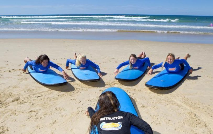 Four people on blue surfboards lying on a sandy beach, facing a surf instructor near the ocean.