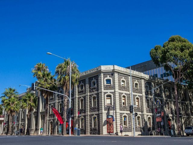 Historic stone building with arched windows and palm trees on a sunny street under a clear blue sky