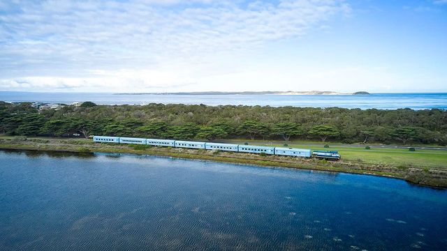 Train crossing a lush island beside a blue river, with ocean and sky in the background