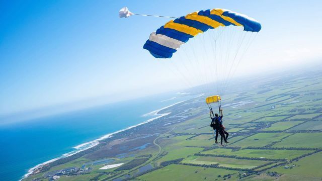 Two skydivers under blue-and-yellow parachutes above a coastal landscape and ocean