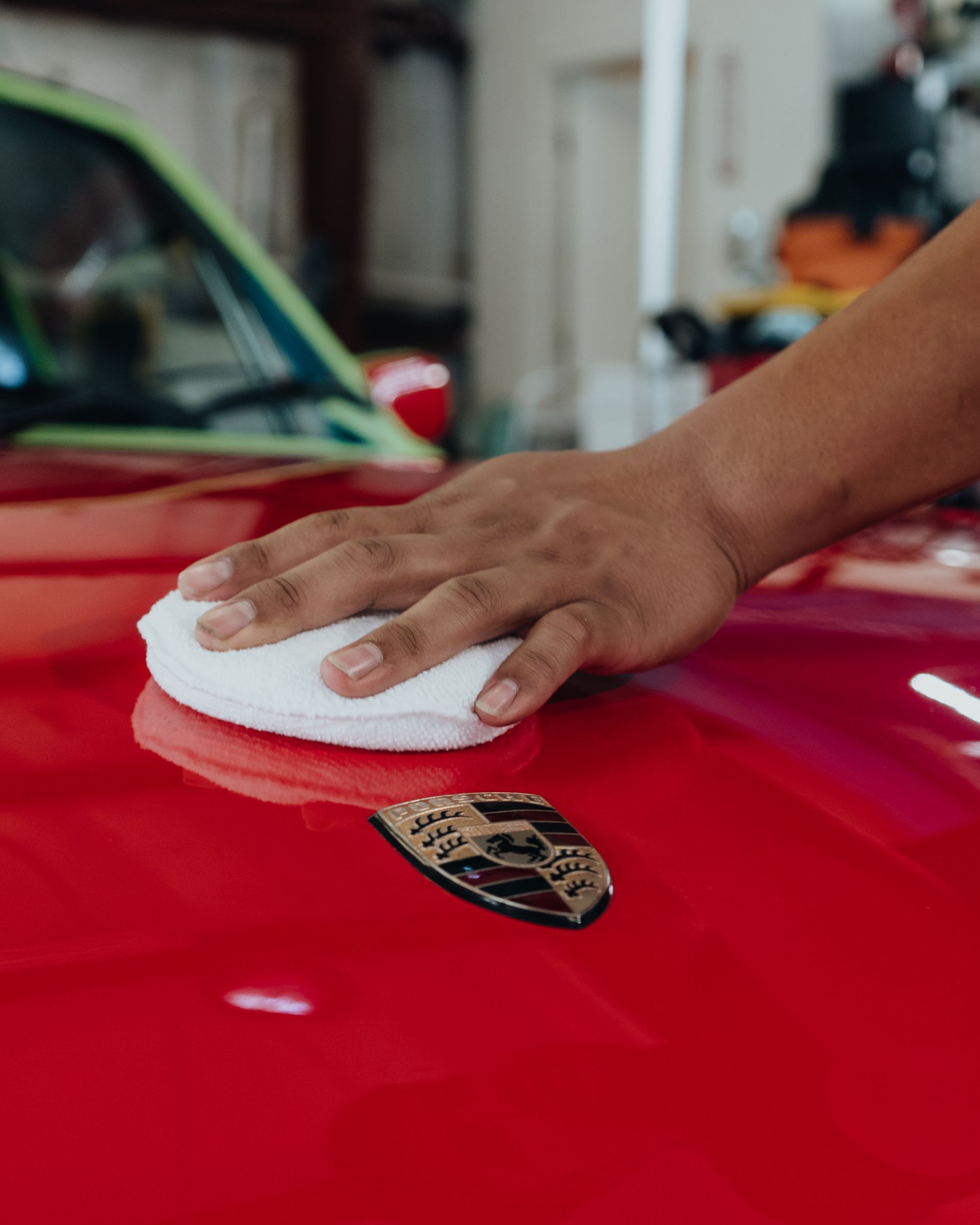 Person's hand polishing red Porsche hood with a white pad, close-up.