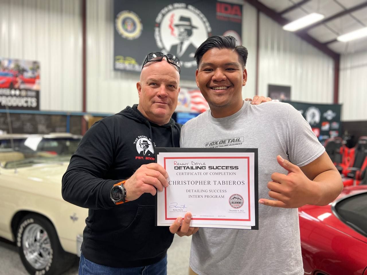 Two men holding a certificate in a garage, smiling. The certificate says