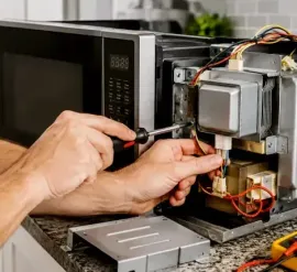 Hands repairing an open microwave on a kitchen counter with tools nearby.