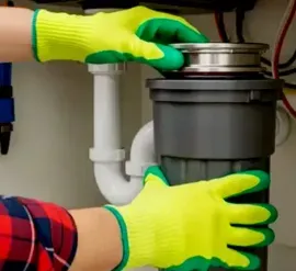 Gloved hands installing a gray garbage disposal under a kitchen sink with pipes visible