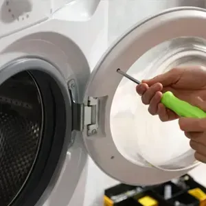 Hands using a green-handled screwdriver to repair a washing machine door hinge