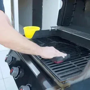 Person cleaning a black gas grill with a cloth, yellow spray bottle on the side.