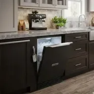 Ice Machine with door open under a dark countertop, beside a sink and coffee maker.