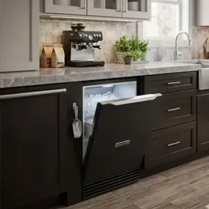 Ice Machine with door open under a dark countertop, beside a sink and coffee maker.